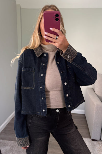 A woman takes a mirror selfie wearing a cropped dark blue denim jacket with leopard-print collar and cuff details, layered over a beige ribbed top and paired with black jeans.