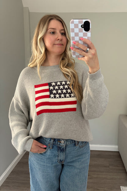 Woman taking a mirror selfie wearing a sweater with an American flag design in a living room.