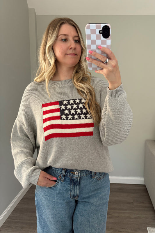 Woman taking a mirror selfie wearing a sweater with an American flag design in a living room.