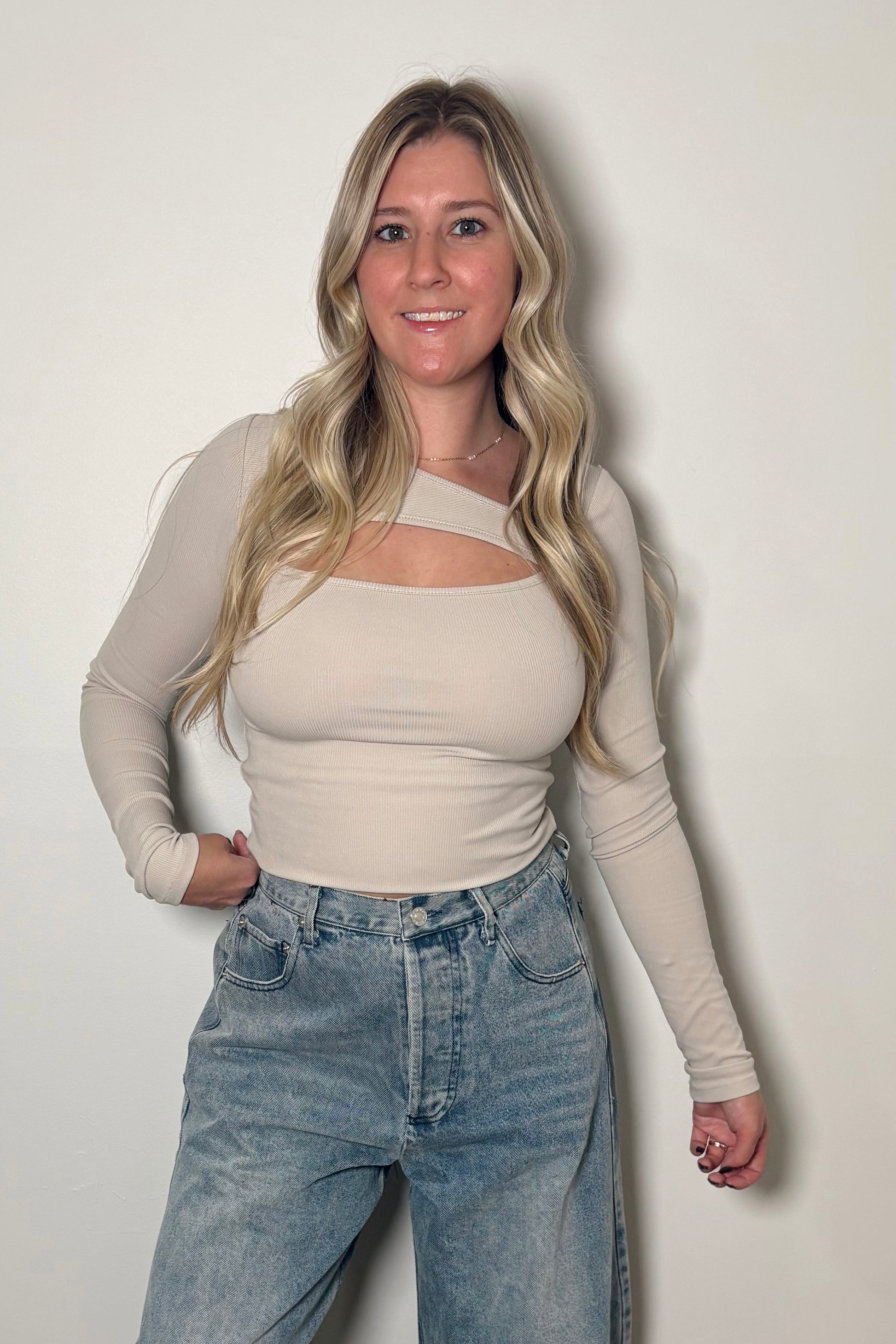 Woman wearing a beige top and blue jeans against a white wall.
