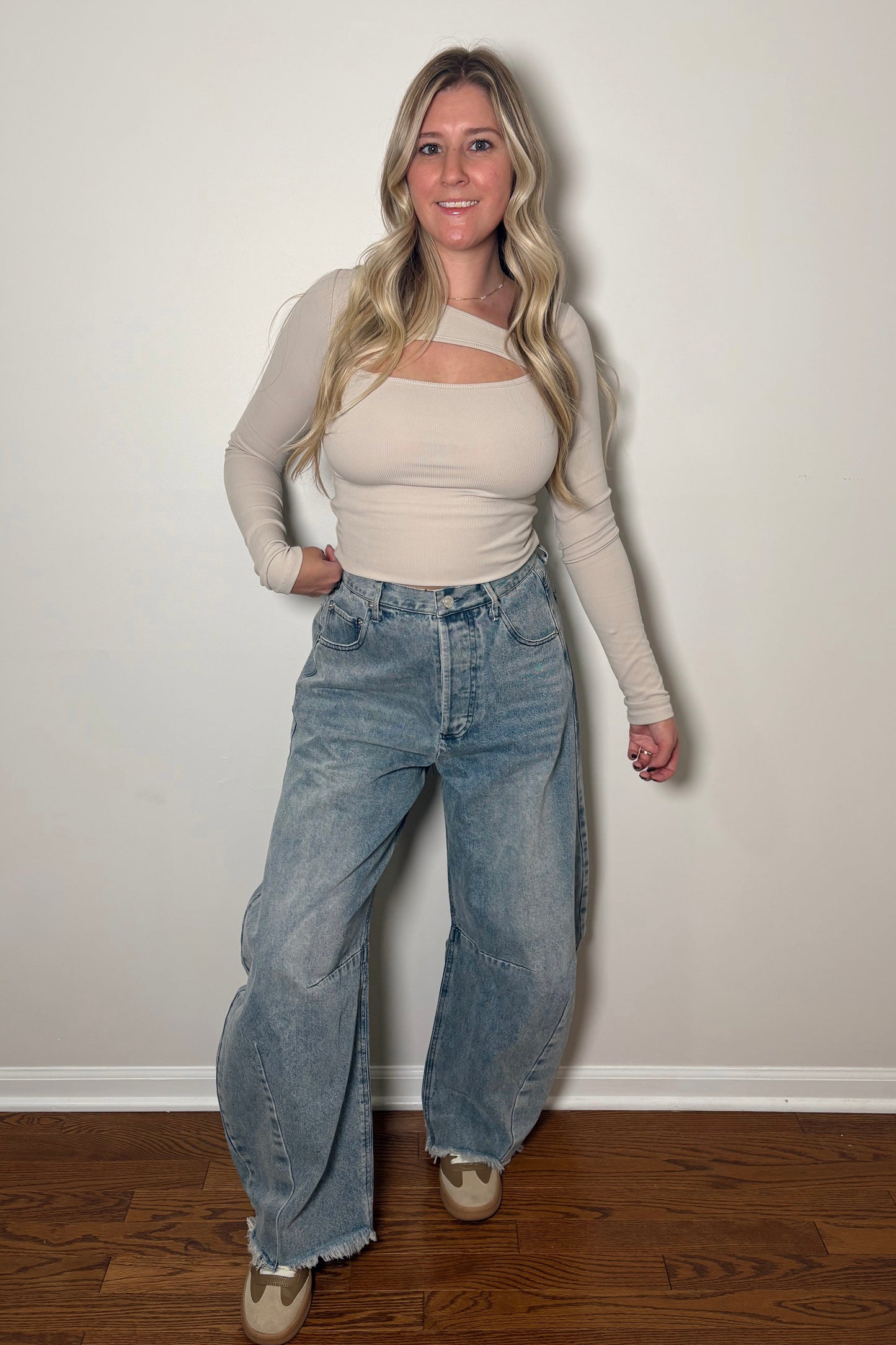 Woman wearing a beige top and blue jeans against a white wall.