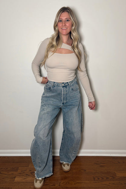 Woman wearing a beige top and blue jeans against a white wall.