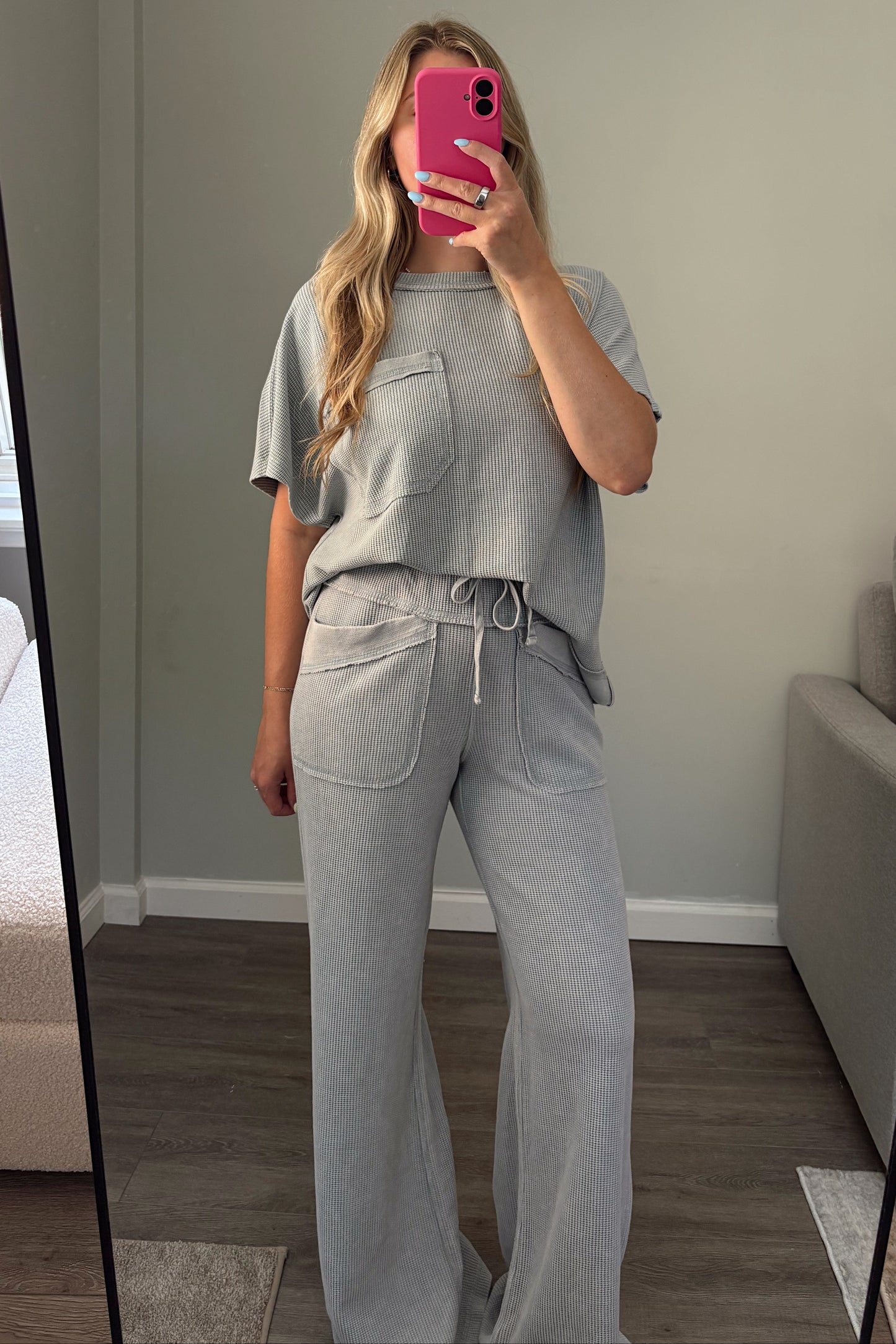 A woman takes a mirror selfie in a room wearing a light blue matching lounge set consisting of a short-sleeve top with chest pockets and high-waisted wide-leg pants with a drawstring waist. She is holding a bright pink smartphone.