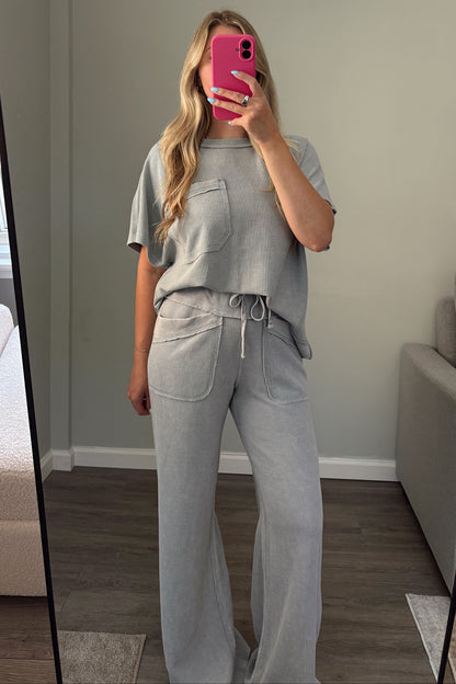 A woman takes a mirror selfie in a room wearing a light blue matching lounge set consisting of a short-sleeve top with chest pockets and high-waisted wide-leg pants with a drawstring waist. She is holding a bright pink smartphone.