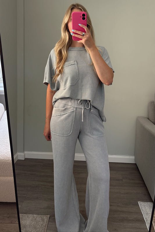 A woman takes a mirror selfie in a room wearing a light blue matching lounge set consisting of a short-sleeve top with chest pockets and high-waisted wide-leg pants with a drawstring waist. She is holding a bright pink smartphone.