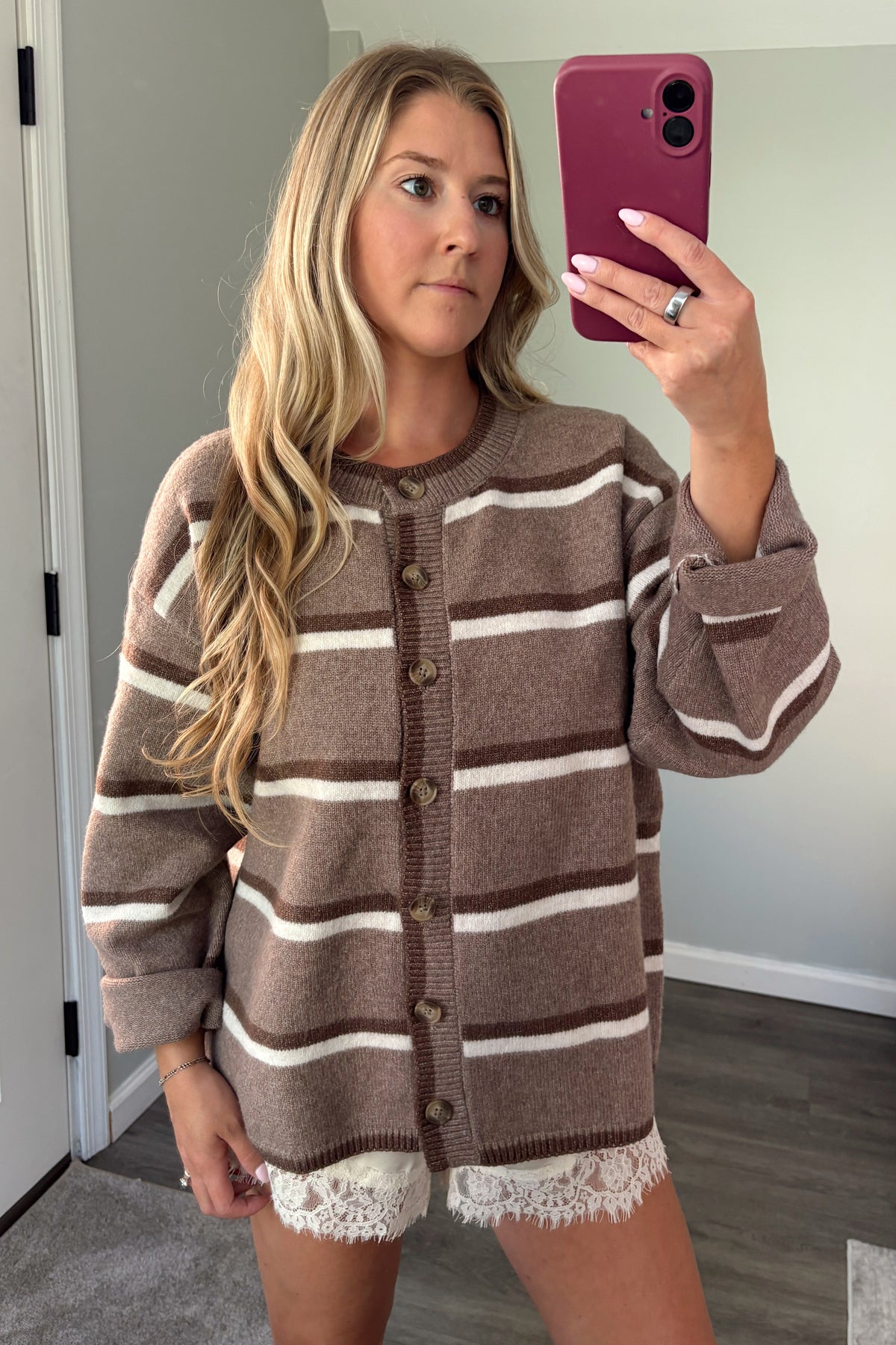 A woman takes a mirror selfie wearing an oversized brown and cream striped knit cardigan with a lace hem, in a home setting.