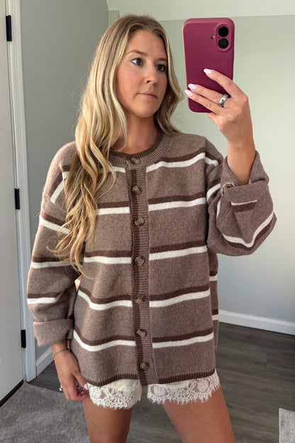 A woman takes a mirror selfie wearing an oversized brown and cream striped knit cardigan with a lace hem, in a home setting.