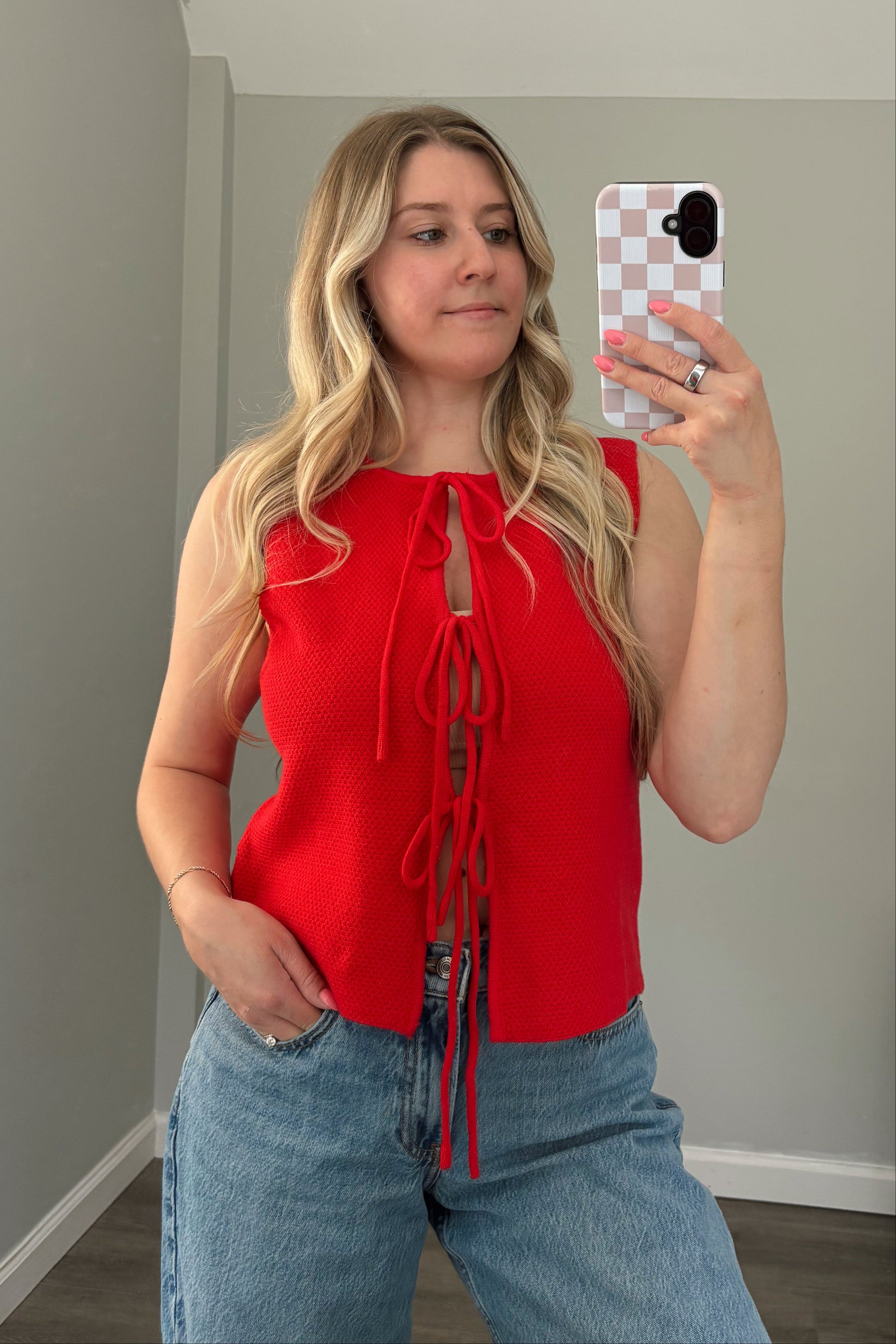 Woman taking a mirror selfie wearing a red top and blue jeans in a room with a window.