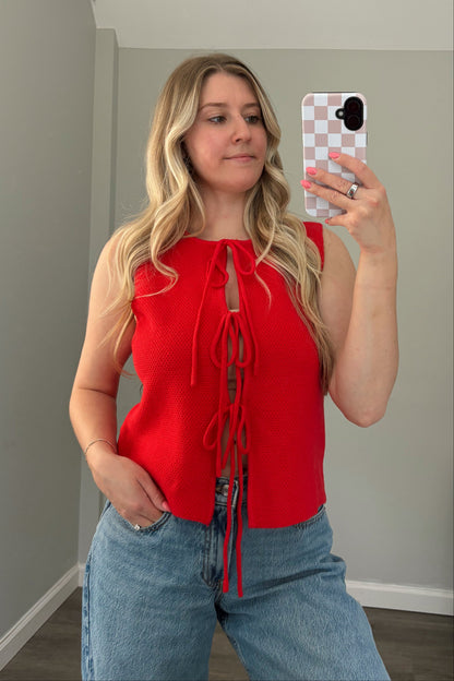 Woman taking a mirror selfie wearing a red top and blue jeans in a room with a window.