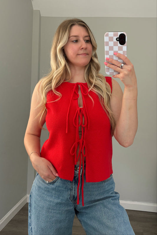 Woman taking a mirror selfie wearing a red top and blue jeans in a room with a window.