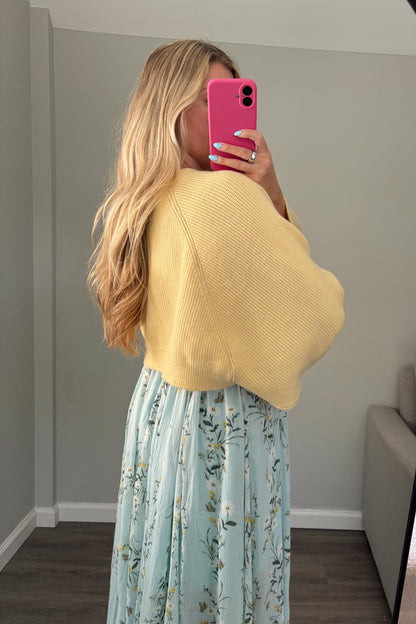 Woman taking a mirror selfie wearing a yellow sweater and light blue floral dress in a room with a couch and window.