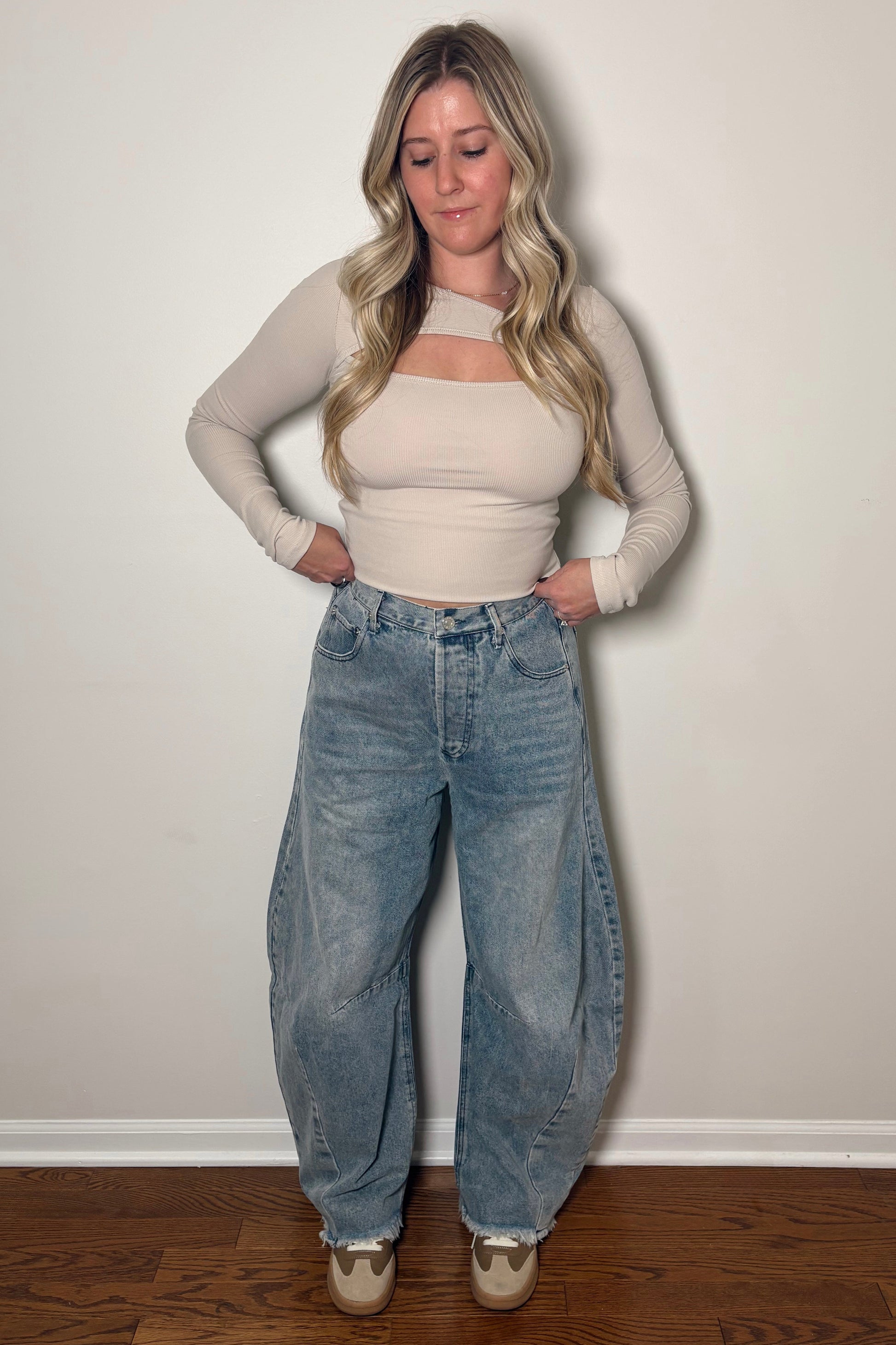 Woman wearing a beige top and blue jeans standing against a plain white wall.