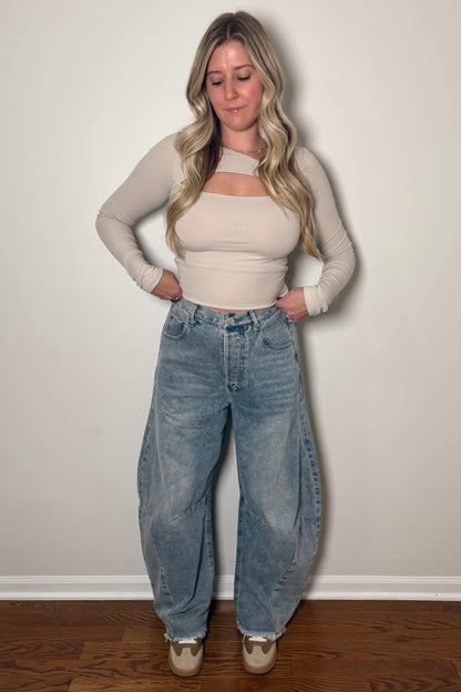 Woman wearing a beige top and blue jeans standing against a plain white wall.