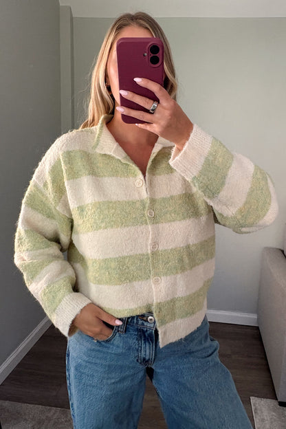 A woman takes a mirror selfie wearing a fuzzy, cropped button-up cardigan with light green and cream horizontal stripes, paired with blue jeans in a living room setting.