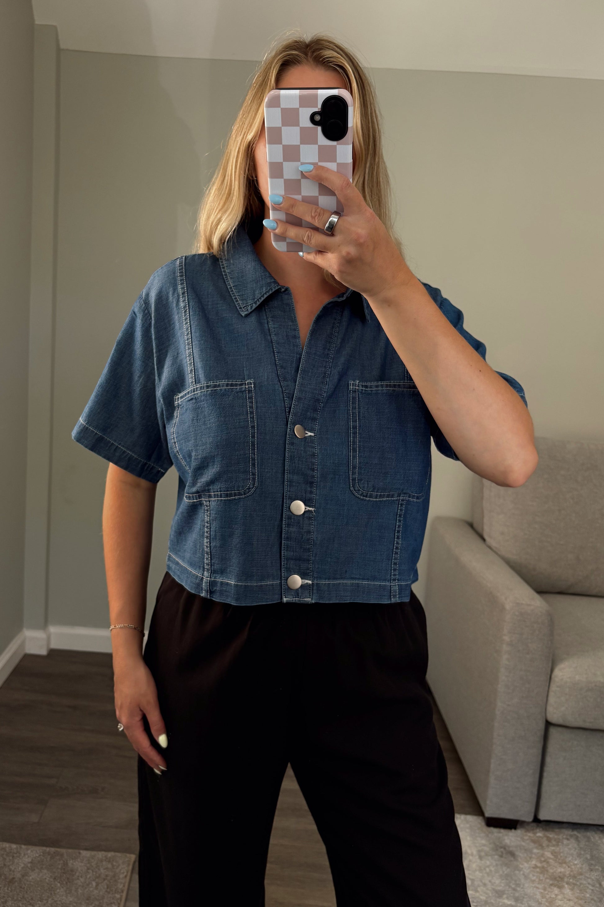 Person wearing a denim shirt taking a mirror selfie in a room with a couch.