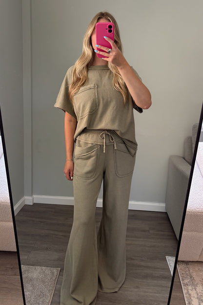 A woman takes a mirror selfie wearing a matching two-piece lounge set in a muted olive green. The set includes a short-sleeve top with a drawstring and matching wide-leg pants.