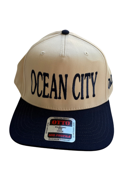 A two-tone snapback cap with a beige crown and a navy blue visor. The front of the cap features the text 'OCEAN CITY' in black, and a vintage-style patch on the brim reads 'OTTO CLASSIC COLDWEAR FIT'.