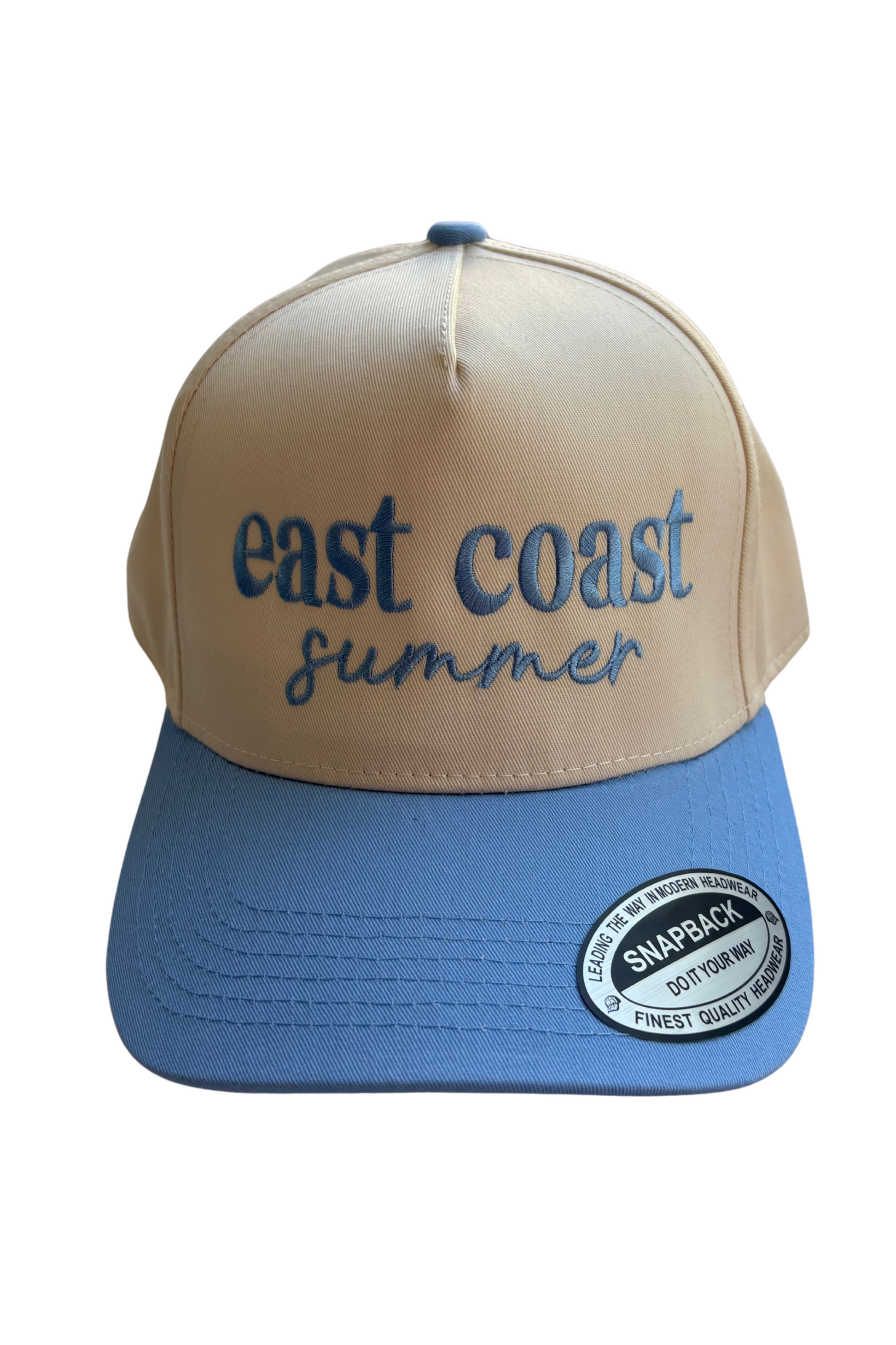 A two-tone trucker hat with a beige front panel and a blue brim. The front panel features the embroidered text 'east coast summer' in blue.