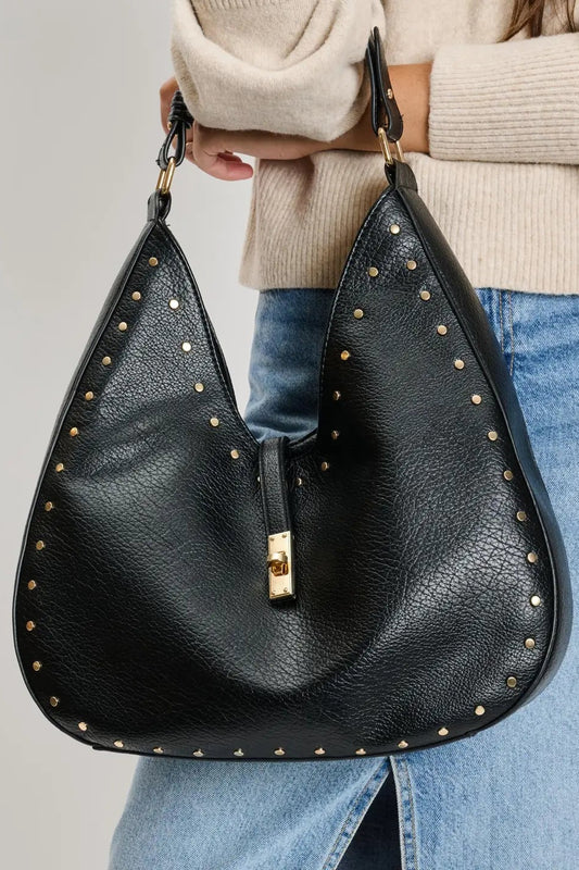 Black studded handbag held by a person wearing a beige sweater and blue jeans on a light gray background
