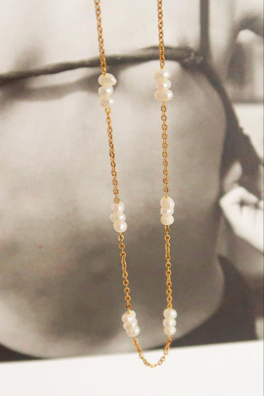 Close-up of the Leilani necklace showing white pearl clusters spaced along a delicate gold-tone chain against a black-and-white photo.

