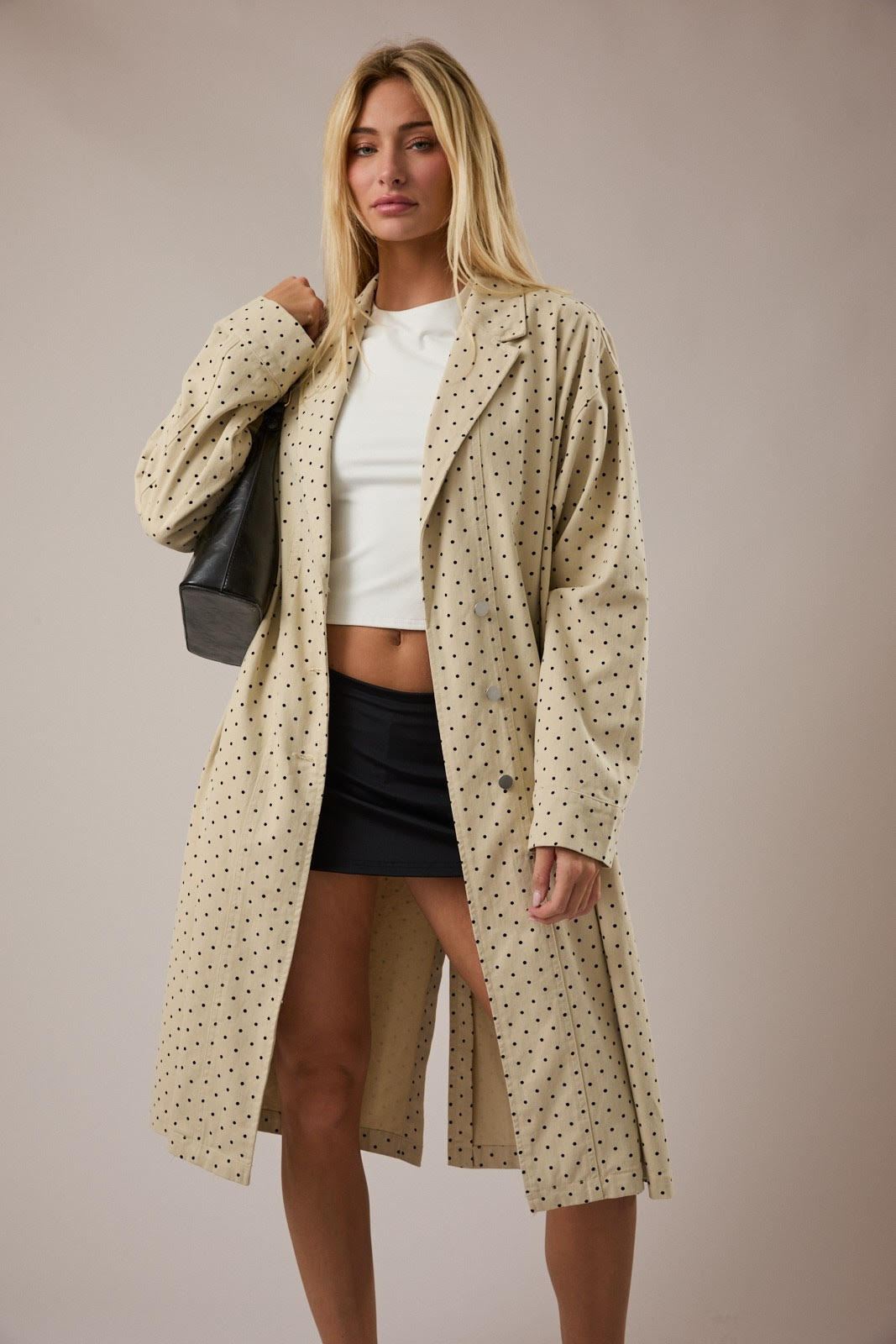 Woman wearing a beige polka dot coat over a white top and black skirt on a plain background
